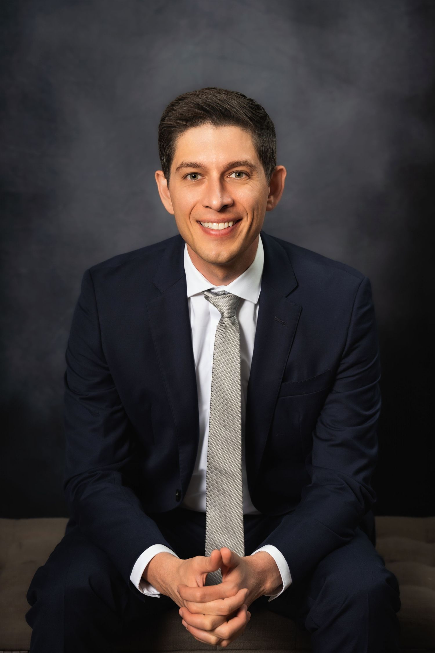 Corporate portrait showing a seated professional in formal attire with clasped hands.