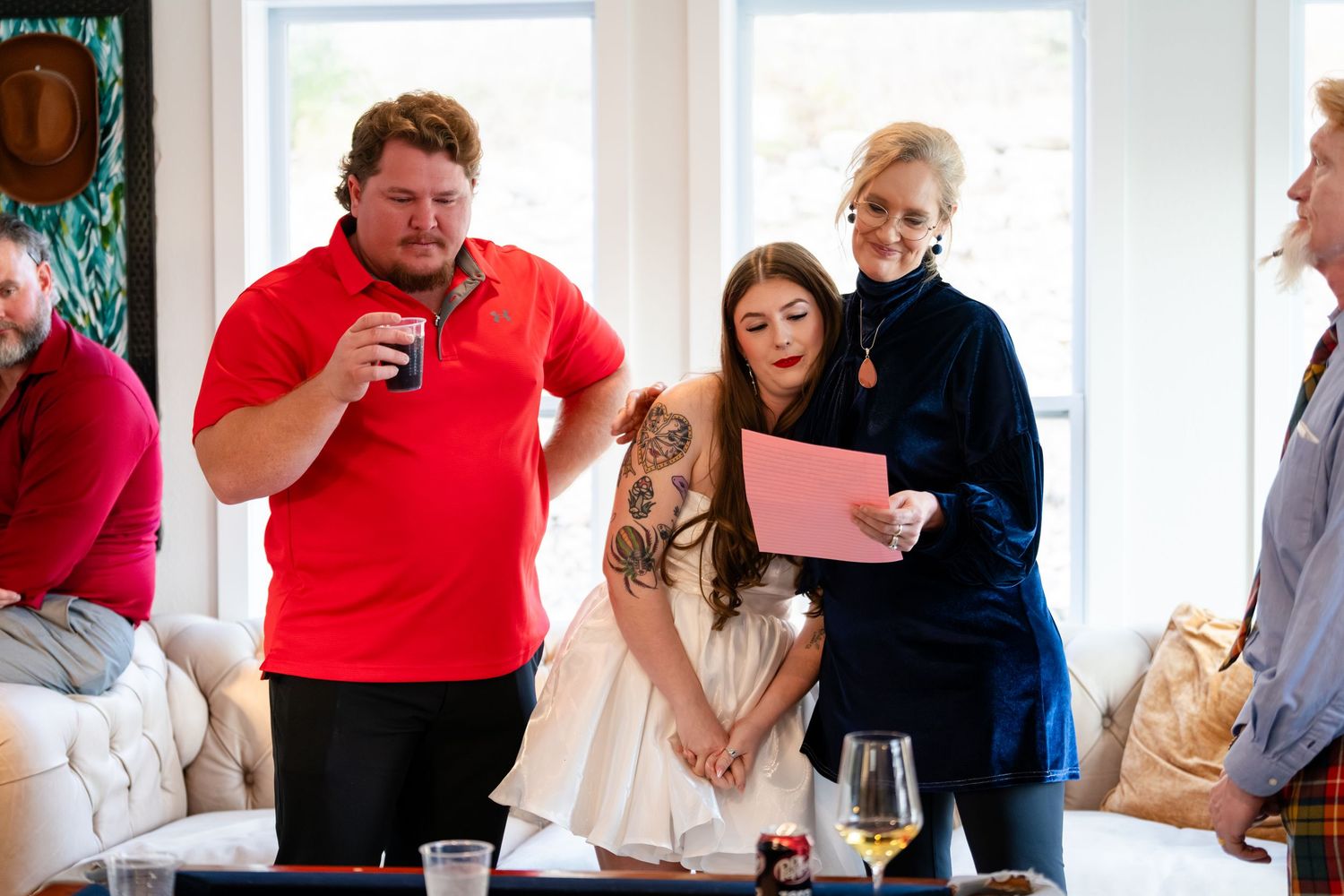 Group of people gathered indoors, woman in white dress reading pink paper while man in red polo holds a drink.