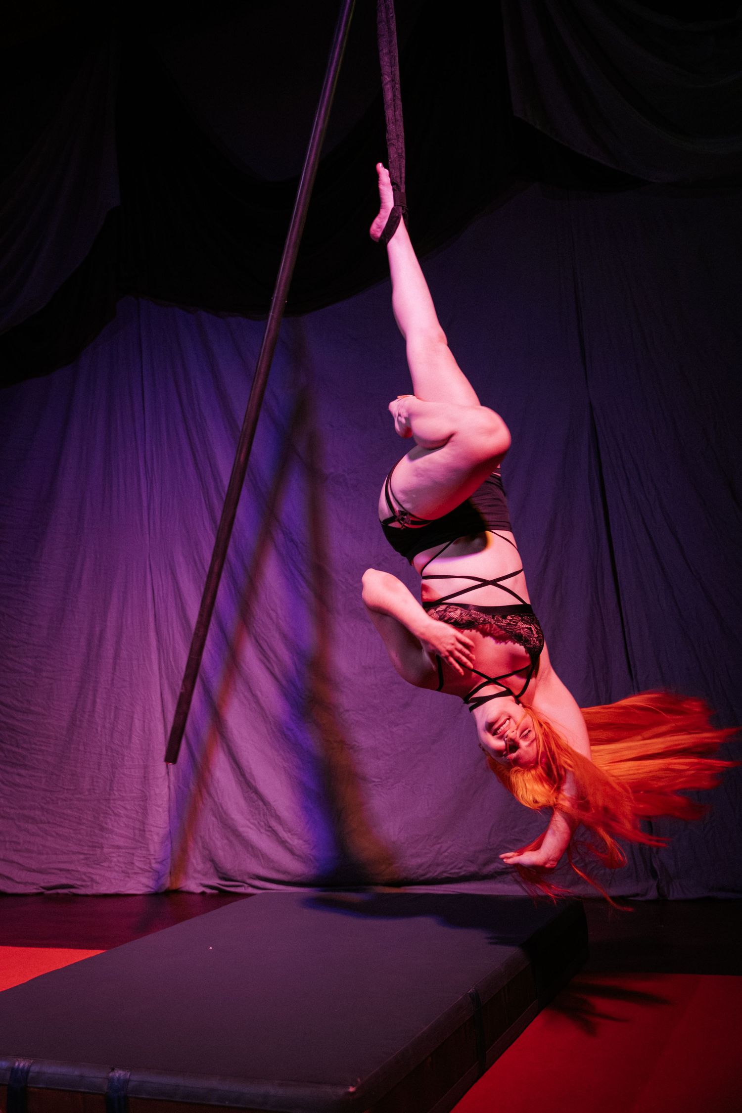Aerial performer in black lingerie wraps around a pole upside down on stage with purple and red lighting.