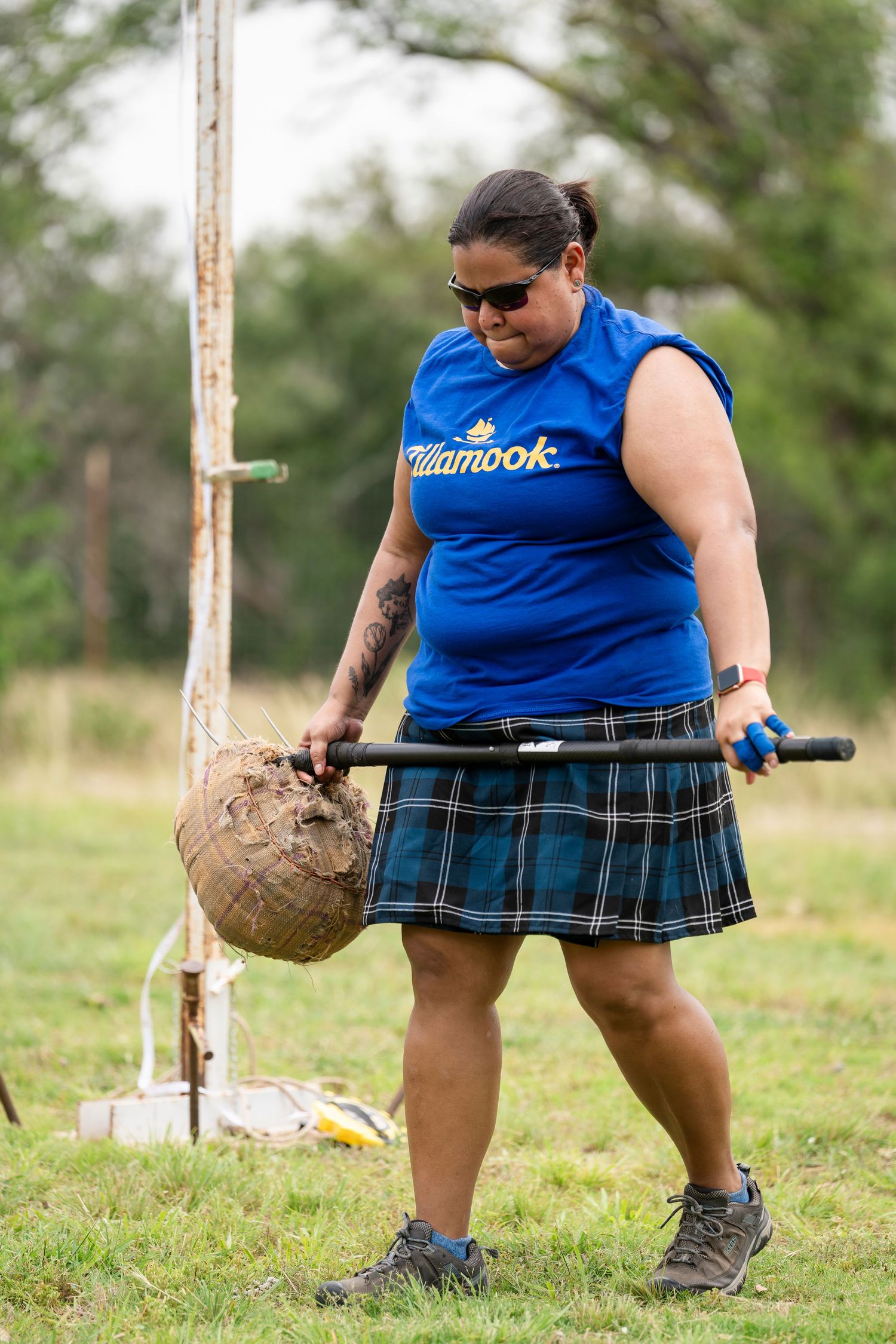 Athlete in blue sleeveless shirt and plaid kilt carries a heavy weight during Highland Games competition.