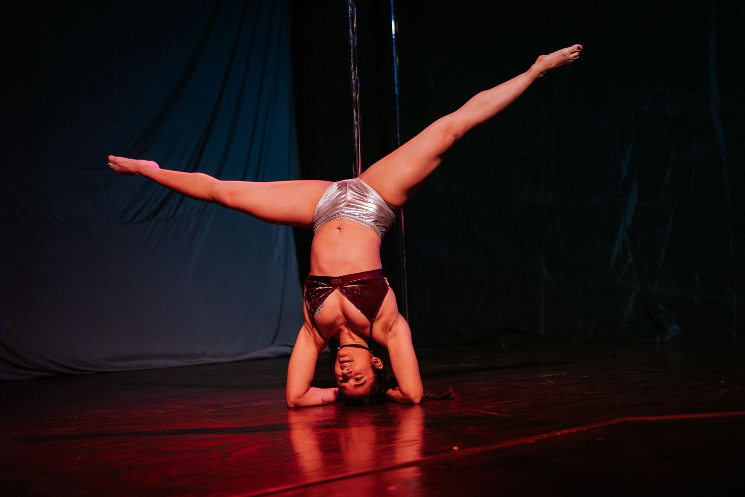 Flexible acrobat performing a headstand split on stage under red lighting in a silver costume.