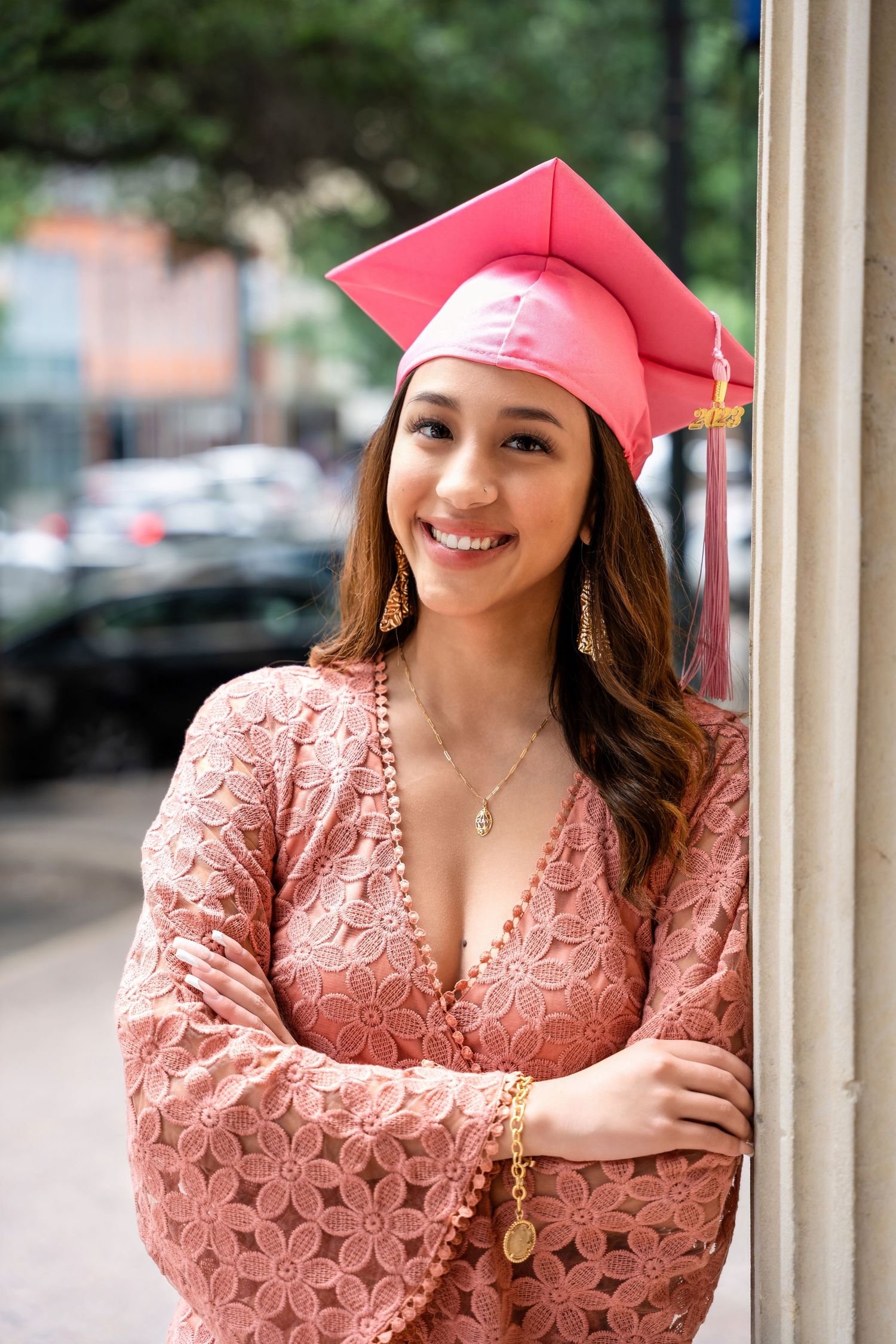 A graduate wearing a pink cap and coral lace dress poses against a stone column.