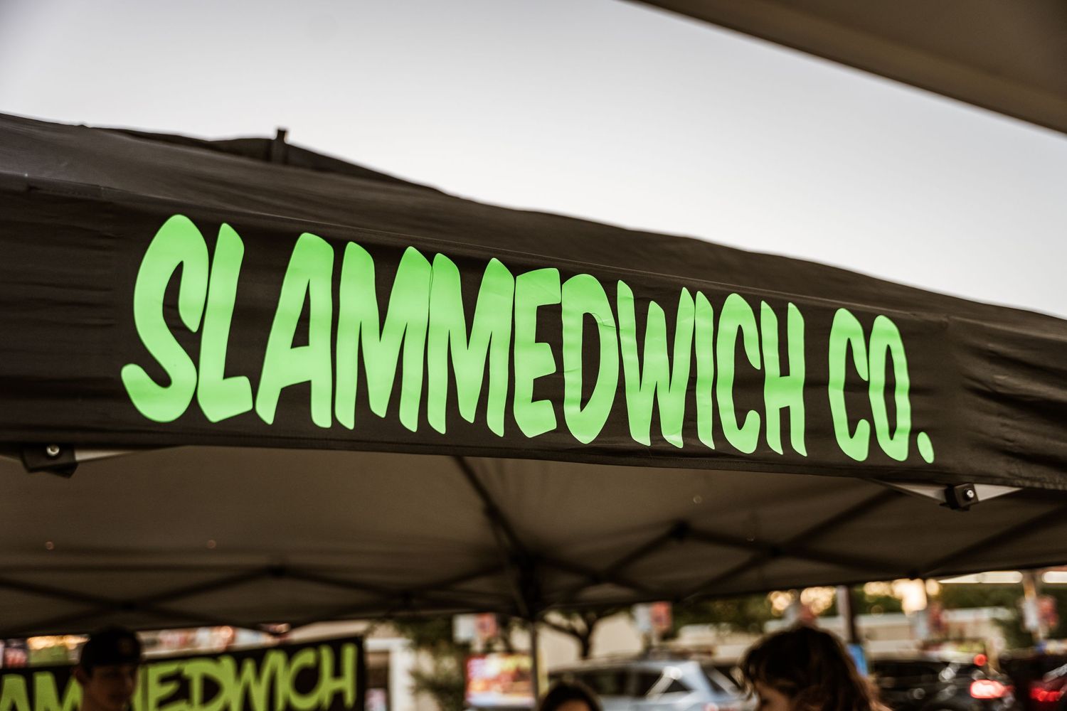Neon green 'SLAMMEDWCH CO.' sign displayed on a dark background at a food vendor booth.