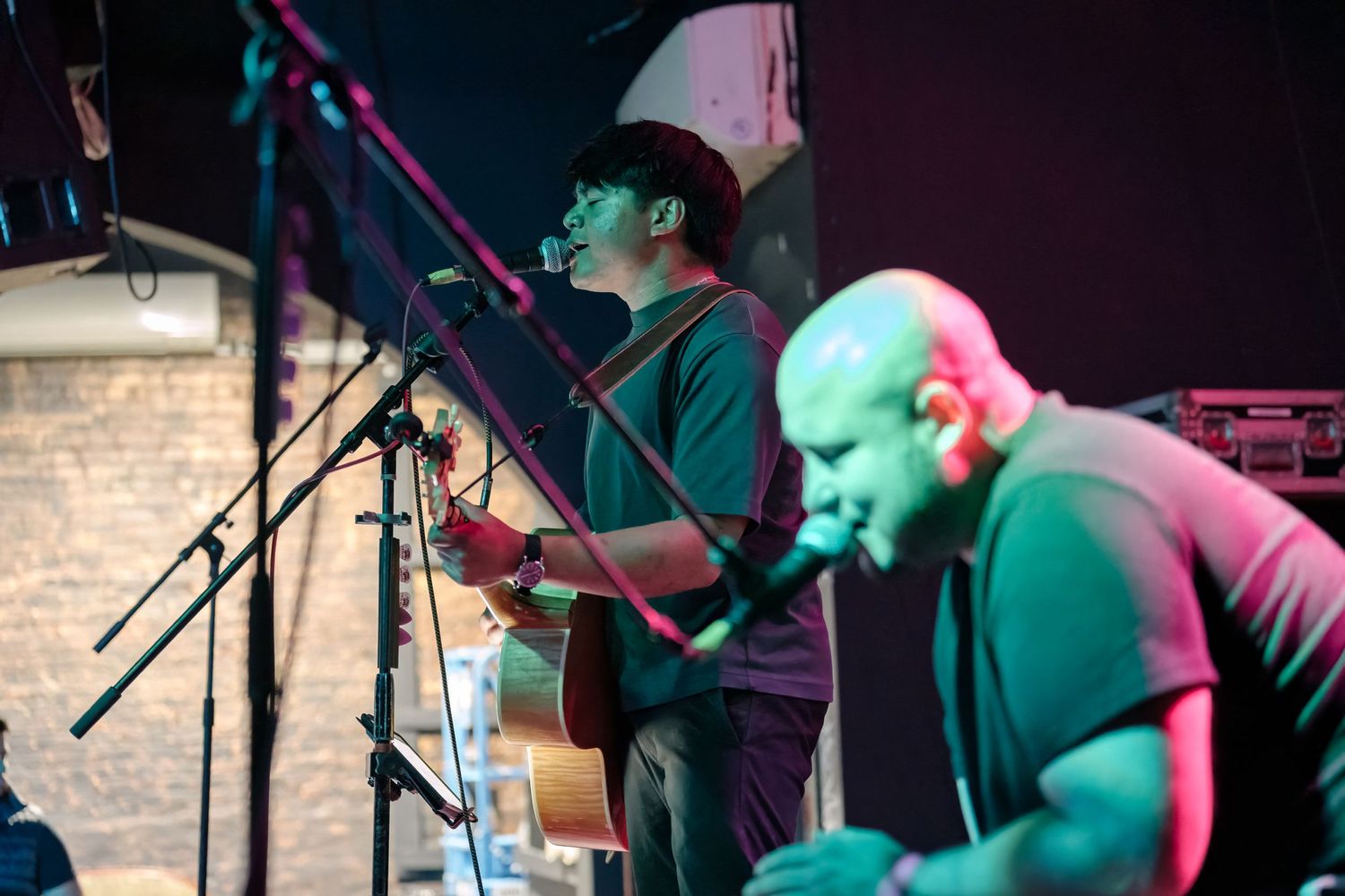 Duo performs acoustic music on stage with dramatic lighting and brick wall backdrop.