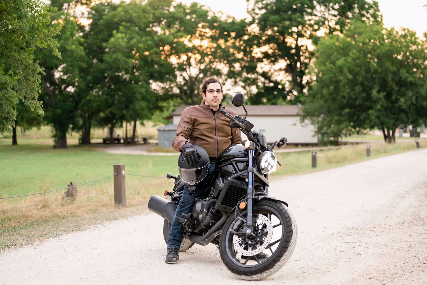 A motorcyclist on a black bike poses on a path with green trees in the background.