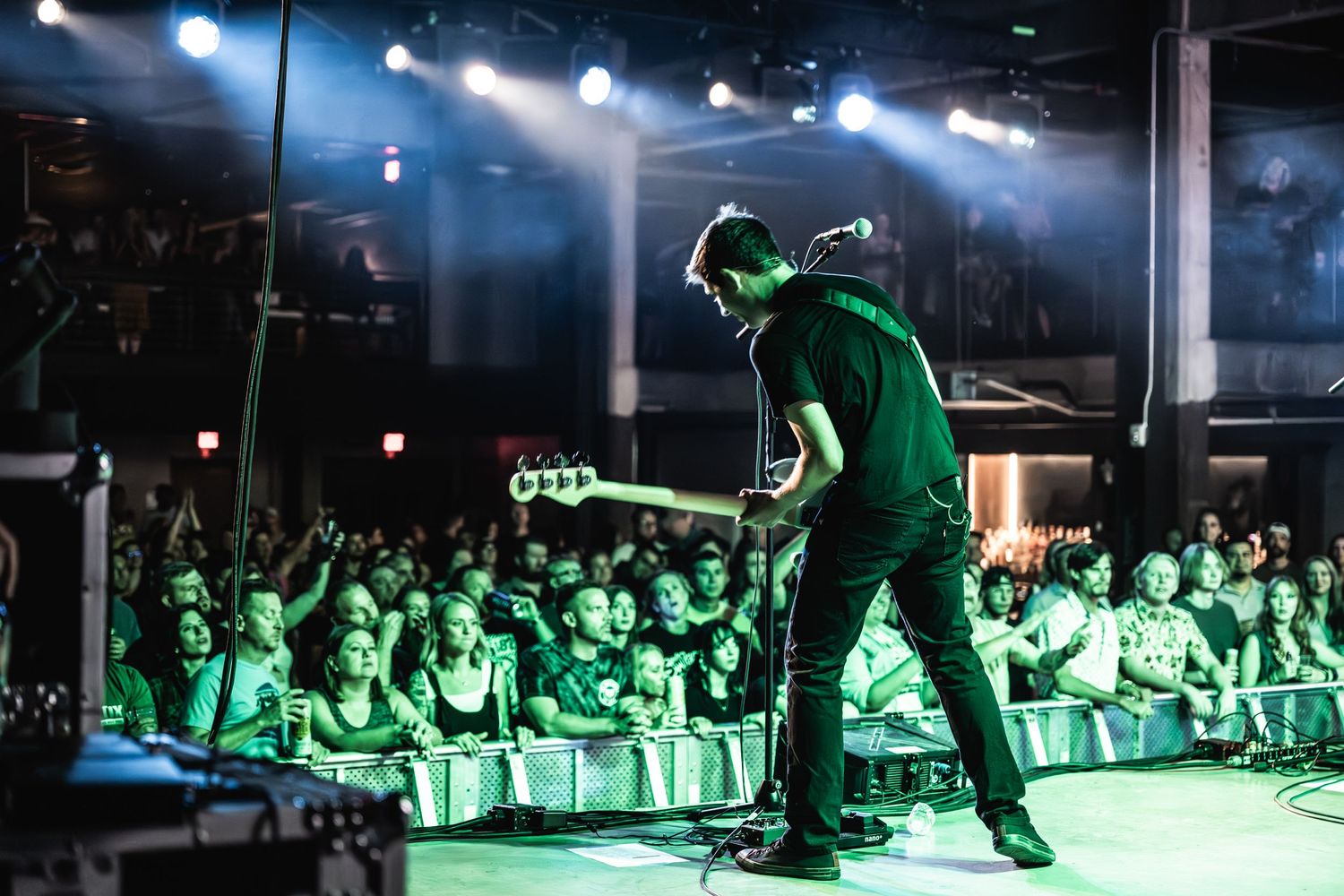 Guitarist silhouetted in green stage lighting performs for packed crowd at indoor venue.