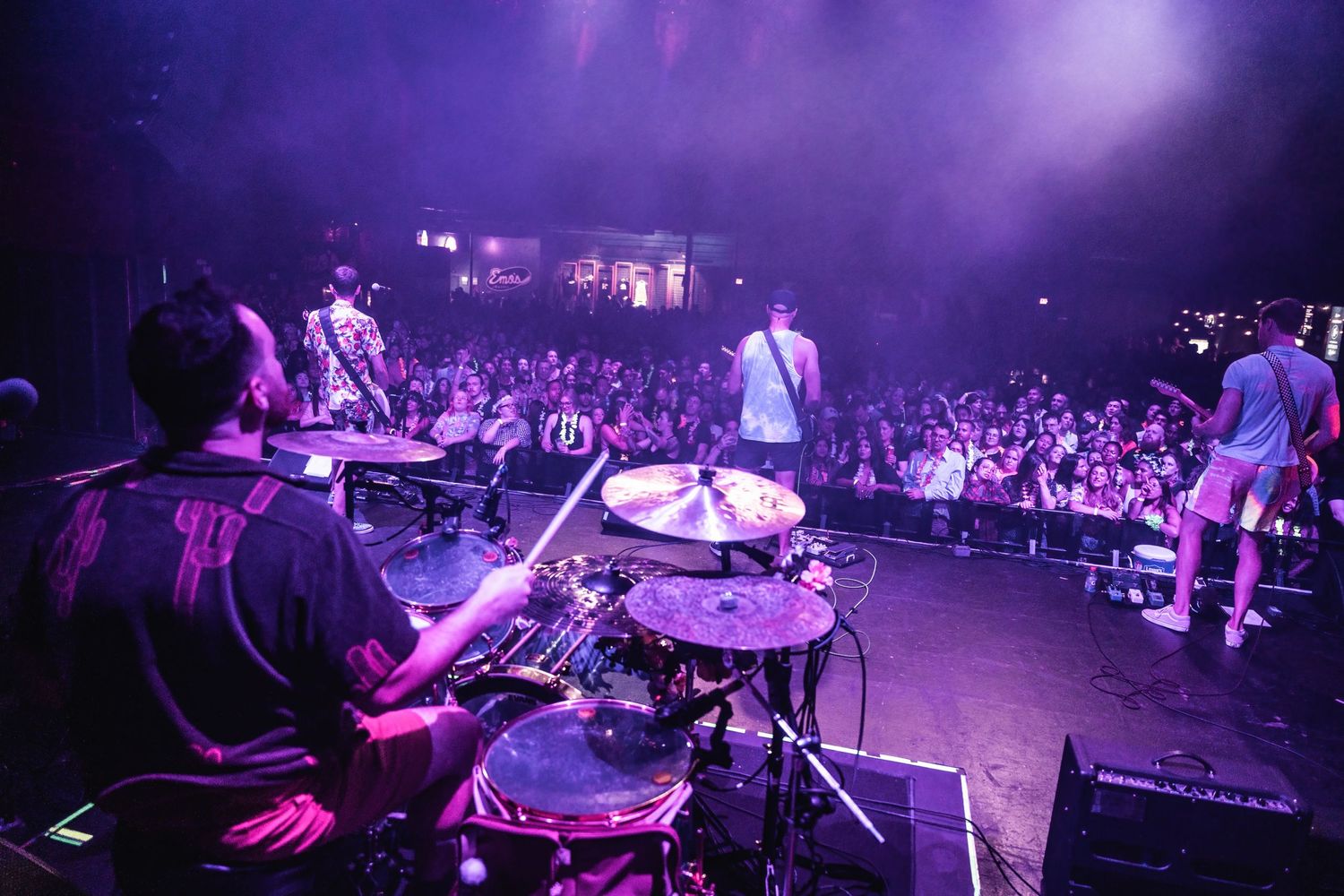 View from drum kit looking out at performing band and crowd under purple stage lights.