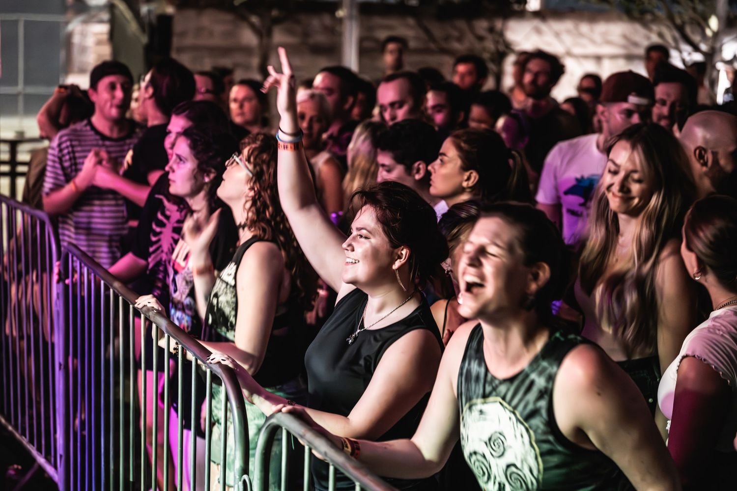 Enthusiastic concert crowd cheers behind safety barrier under purple stage lighting.