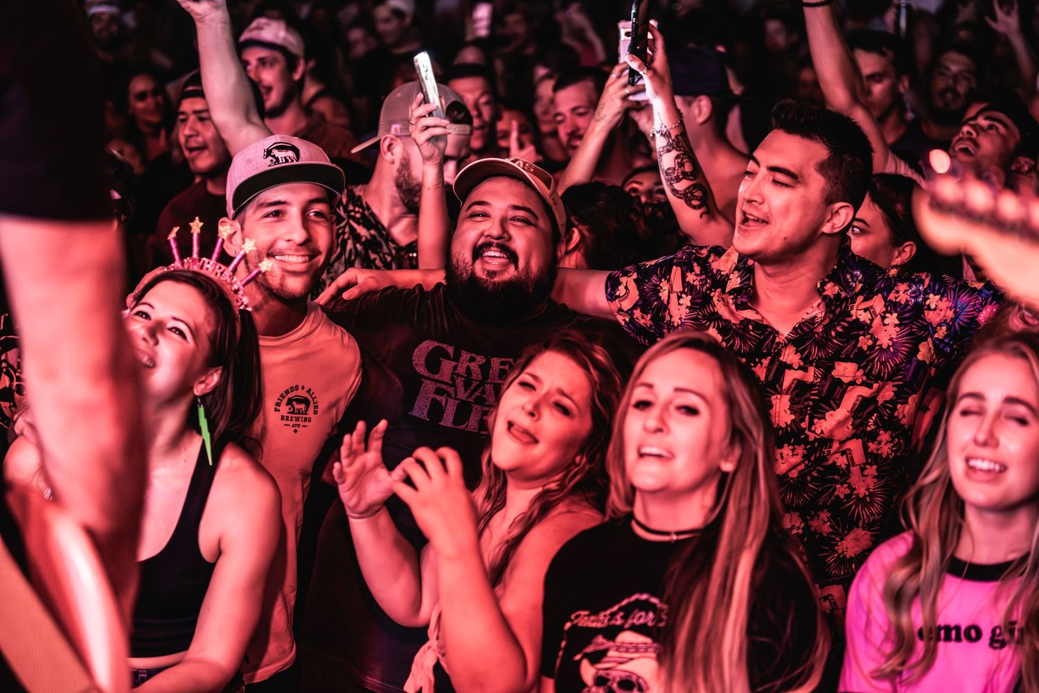 Enthusiastic concert crowd with arms raised bathed in pink lighting at a live music event.