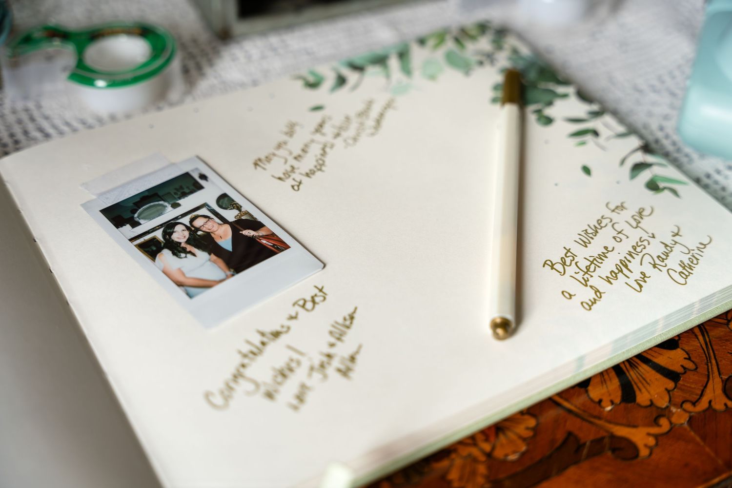 Wedding guestbook with polaroid photo and pen on wooden table with greenery details.
