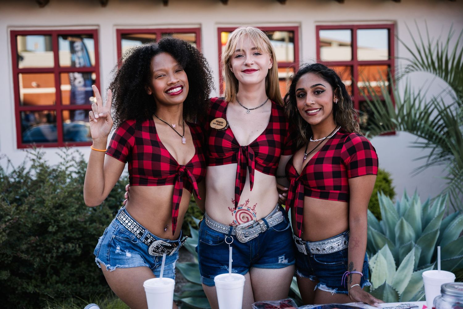 Three friends in matching red plaid crop tops and denim shorts pose together at outdoor gathering.