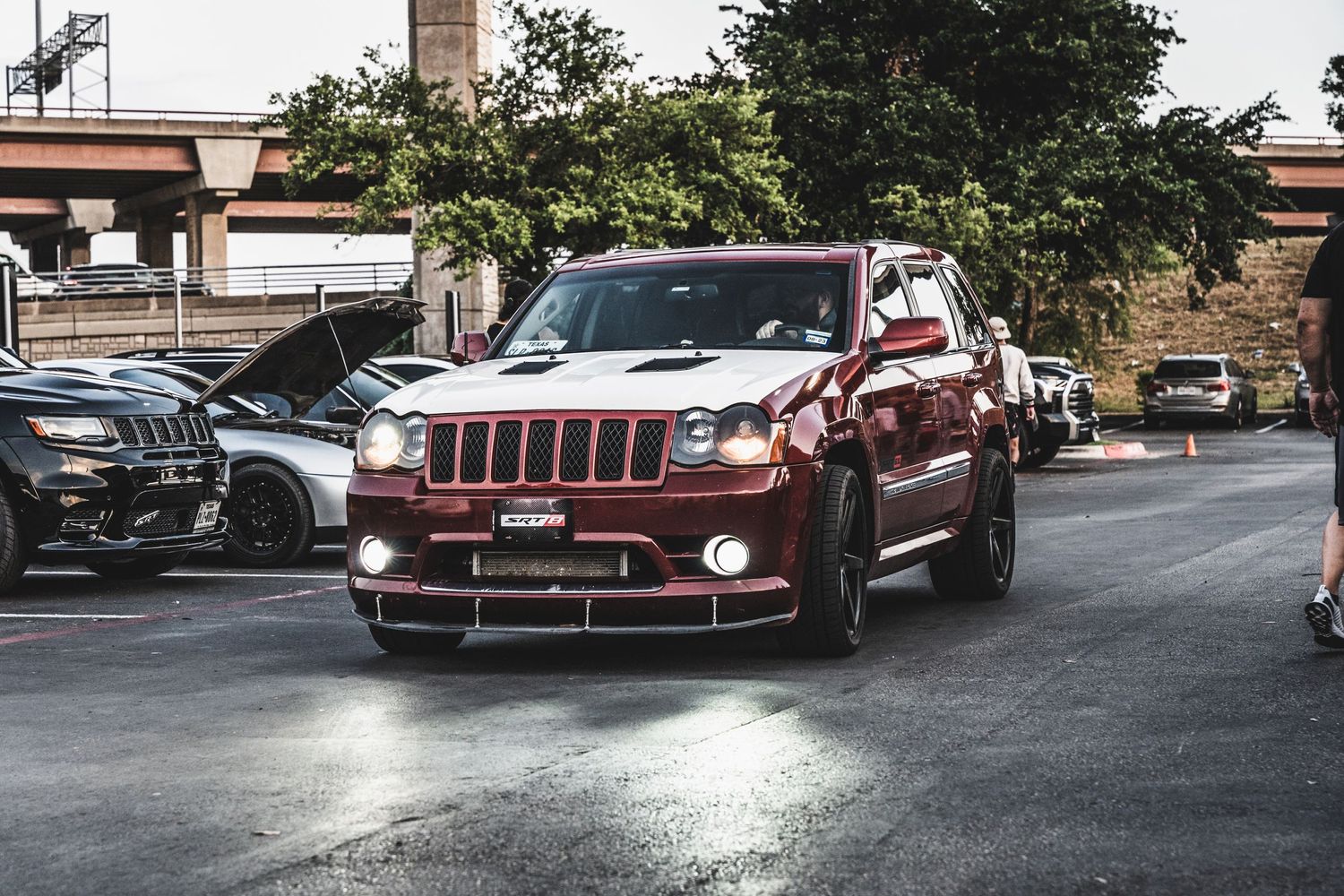 Red Jeep Grand Cherokee driving through a parking lot at a car show.