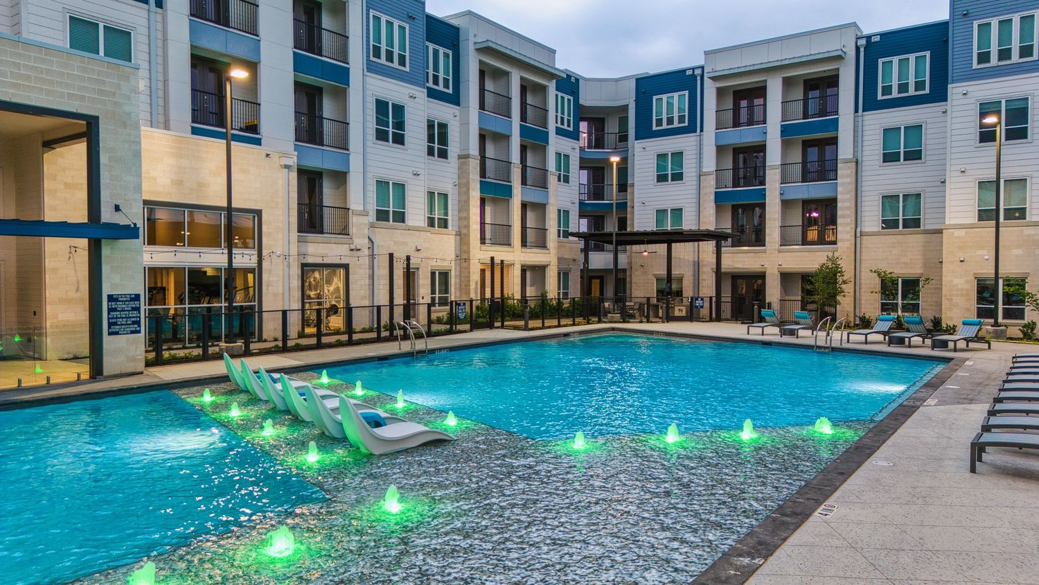 Modern apartment complex featuring illuminated pool with gray deck and landscaping.