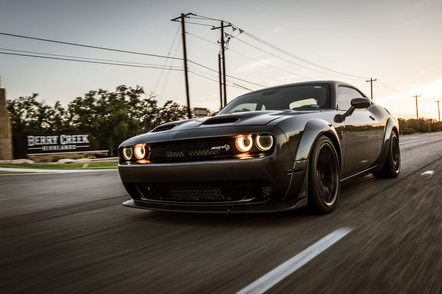 Black Dodge Challenger SRT widebody with glowing headlights speeding on a street at dusk.
