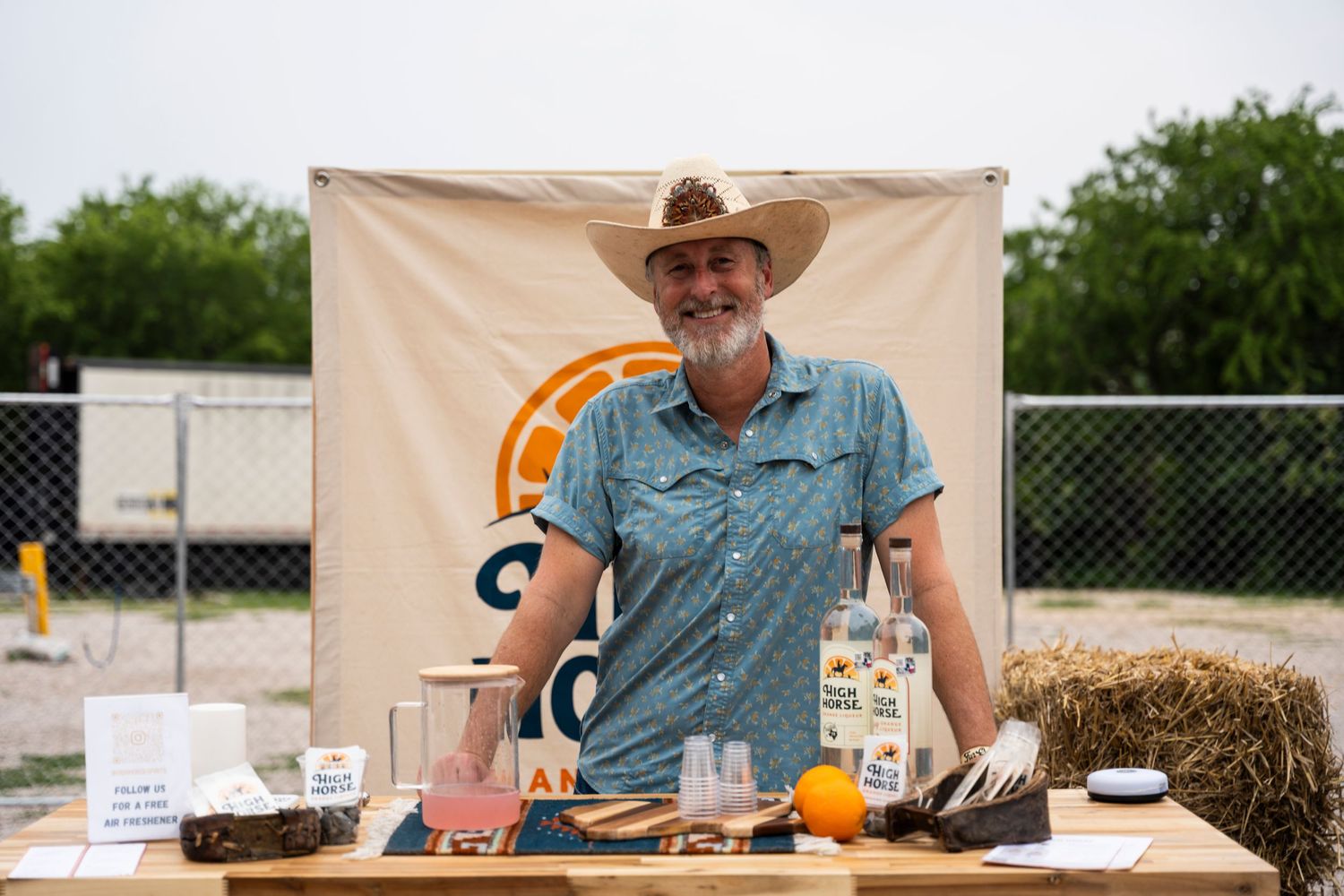 A vendor in a cowboy hat and blue shirt stands behind a display table with hay bales at an outdoor market event.