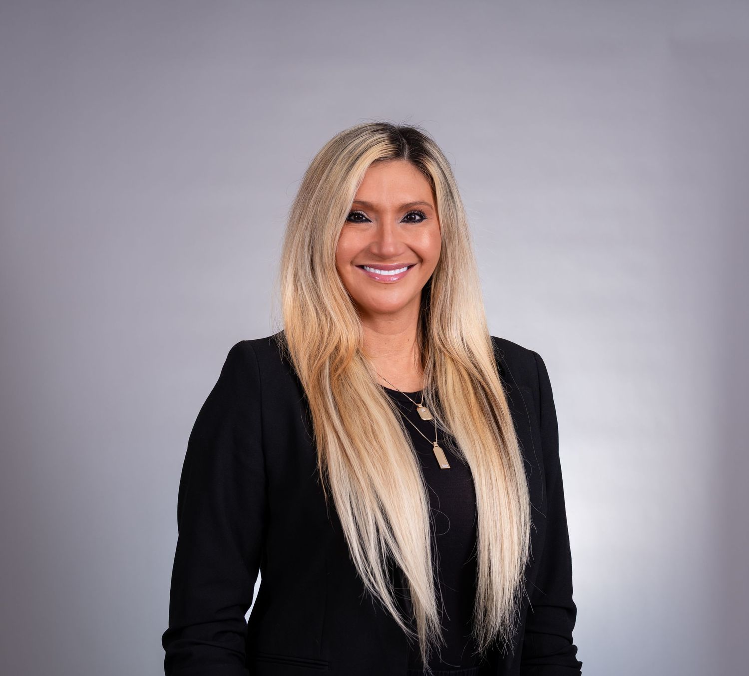 Professional portrait of someone with long blonde hair wearing a black blazer against a gray studio background.