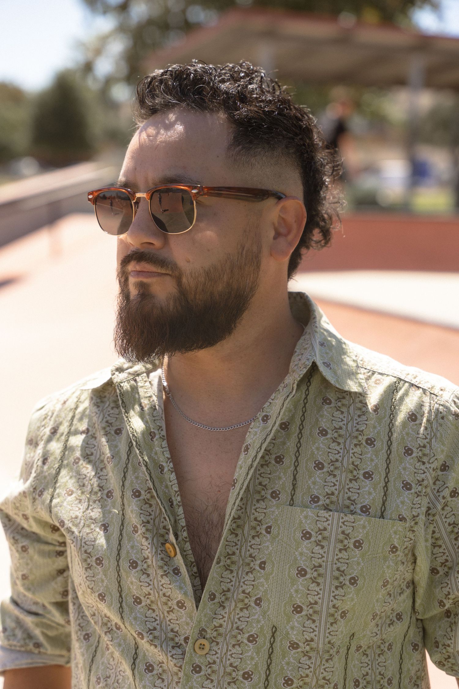 A person wearing sunglasses and a patterned shirt poses in natural lighting outdoors.