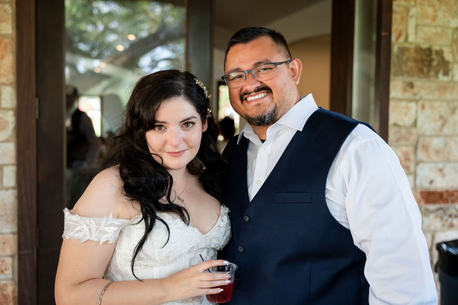 A couple smile together at a wedding reception, with the woman wearing a white off-shoulder dress and man in a navy vest.