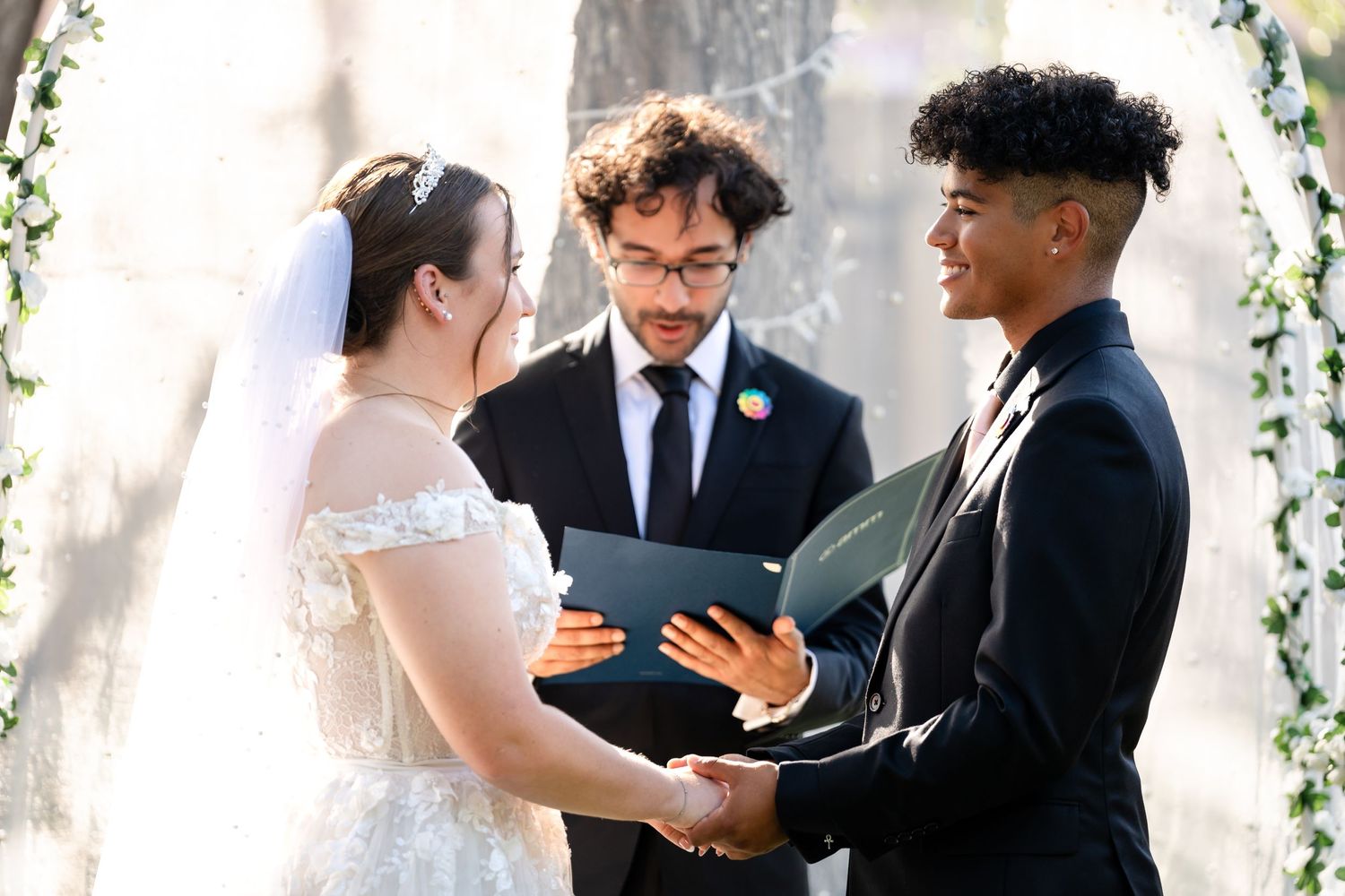 An intimate outdoor wedding ceremony takes place beneath a decorated arch with vines and flowers.