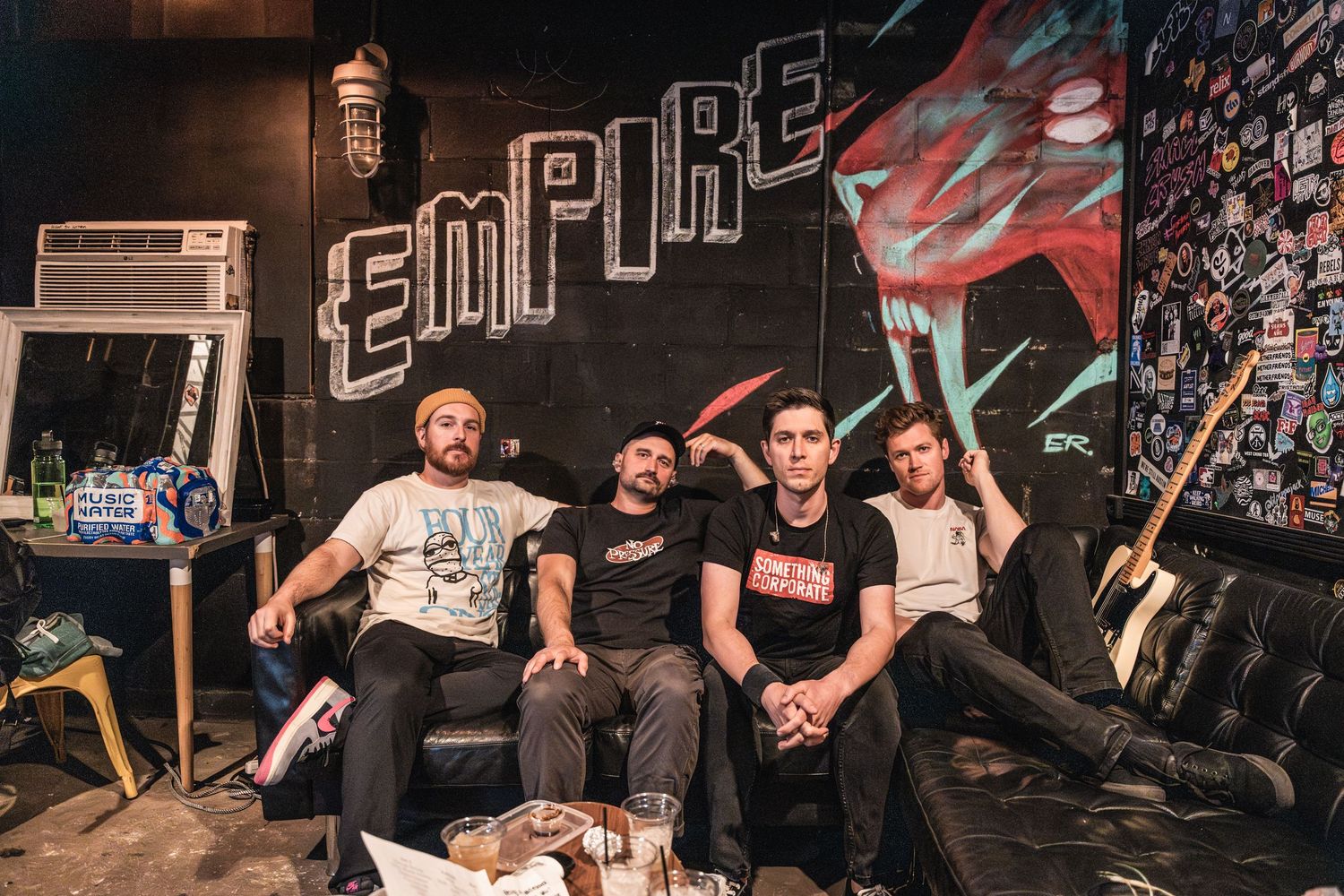 A group of band members pose together on a couch against a wall with street art and the word 'Empire' painted behind them.