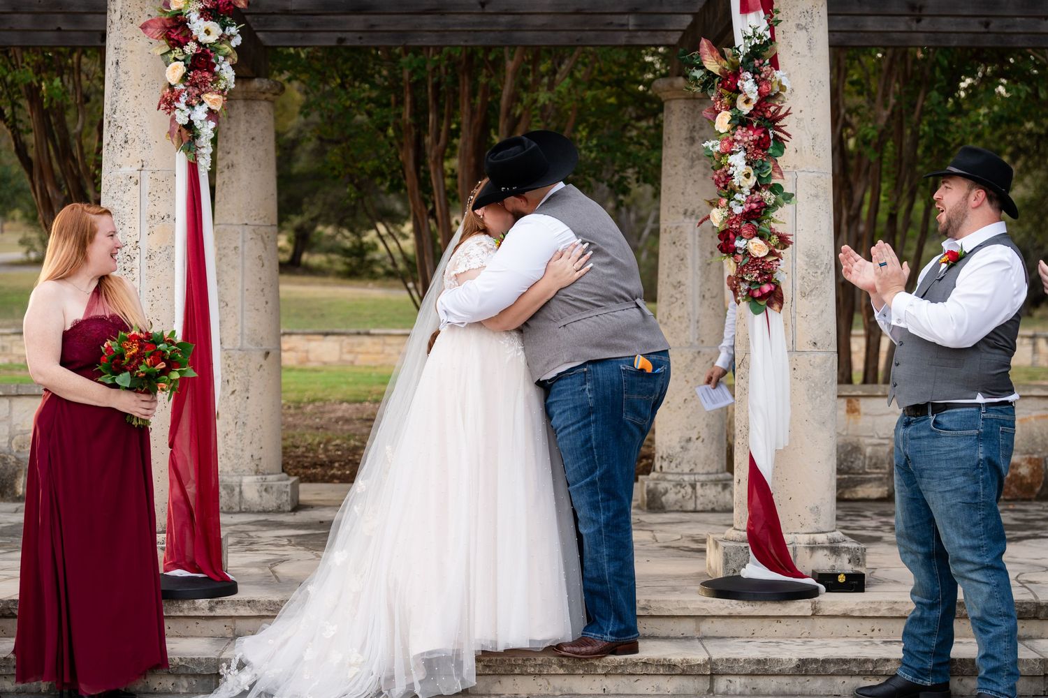 A wedding ceremony kiss under a pergola decorated with burgundy and white floral arrangements.