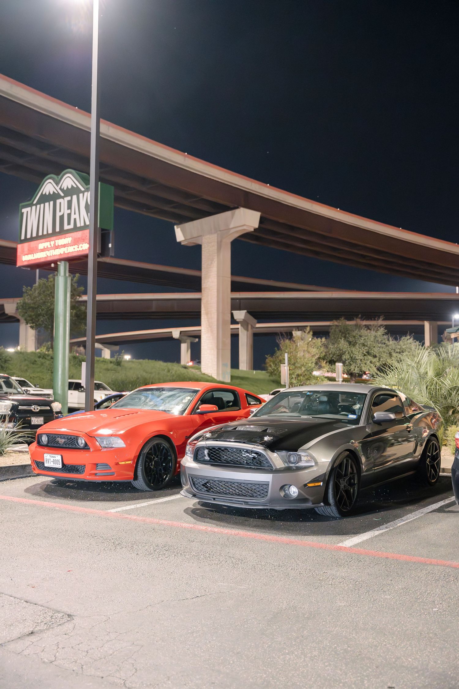 Two Ford Mustangs, one red and one gray, parked under a Twin Peaks restaurant sign at night.