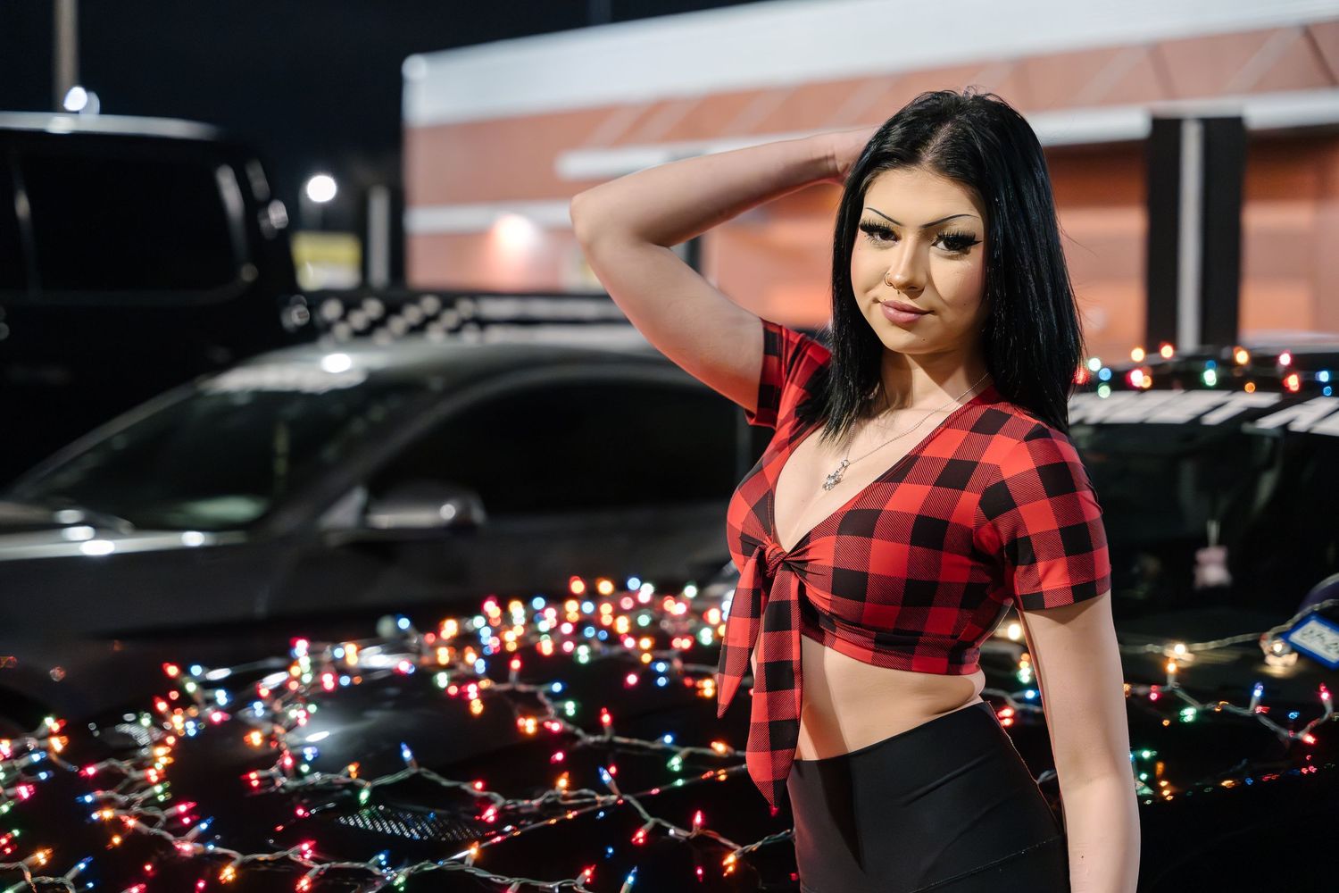 Person in red plaid crop top poses next to car decorated with string lights at night.