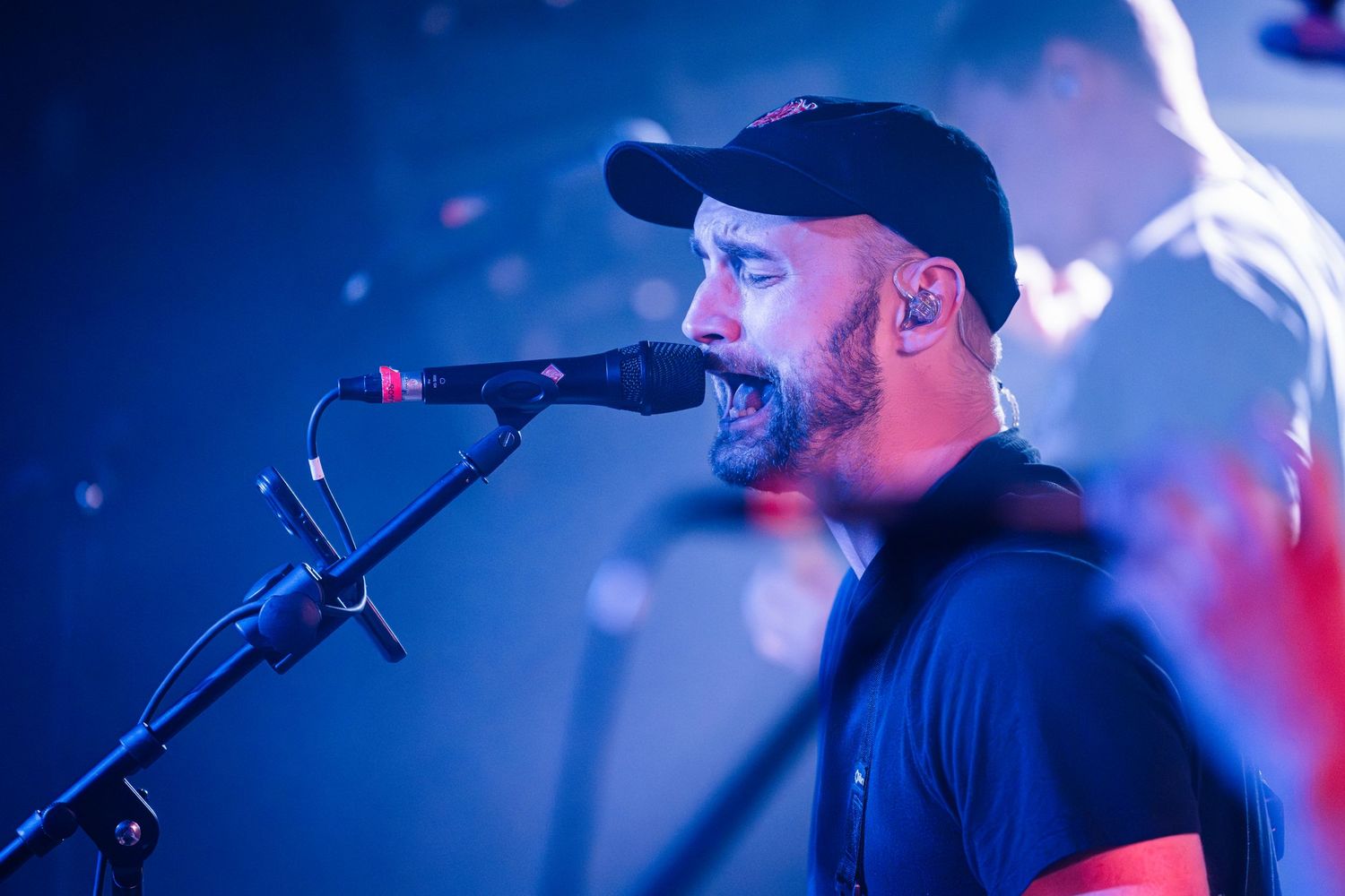 Concert performer with a baseball cap singing into a microphone against blue stage lighting.