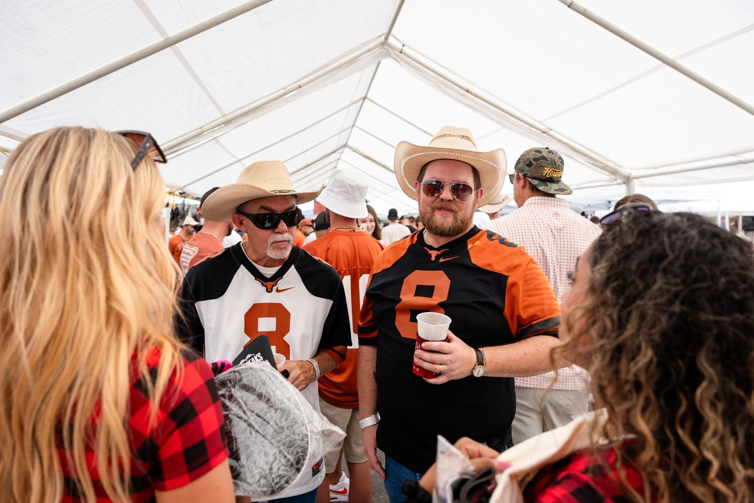 Sports fans in orange jerseys socializing under white tent at tailgate party.