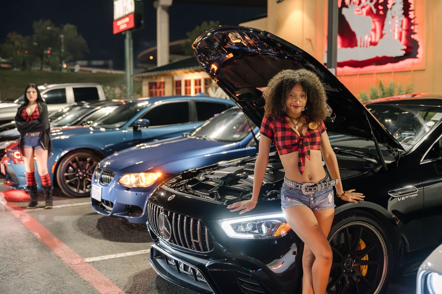 Woman posing by black Mercedes AMG with open hood at night car meet.