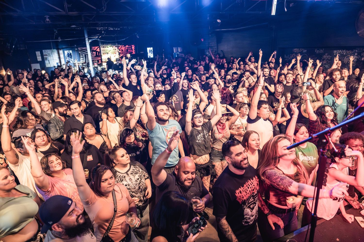 Large crowd with raised hands fills a dark concert venue during an energetic performance.