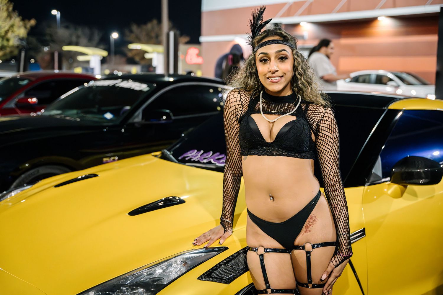 Model in black outfit poses next to yellow sports car at night event.