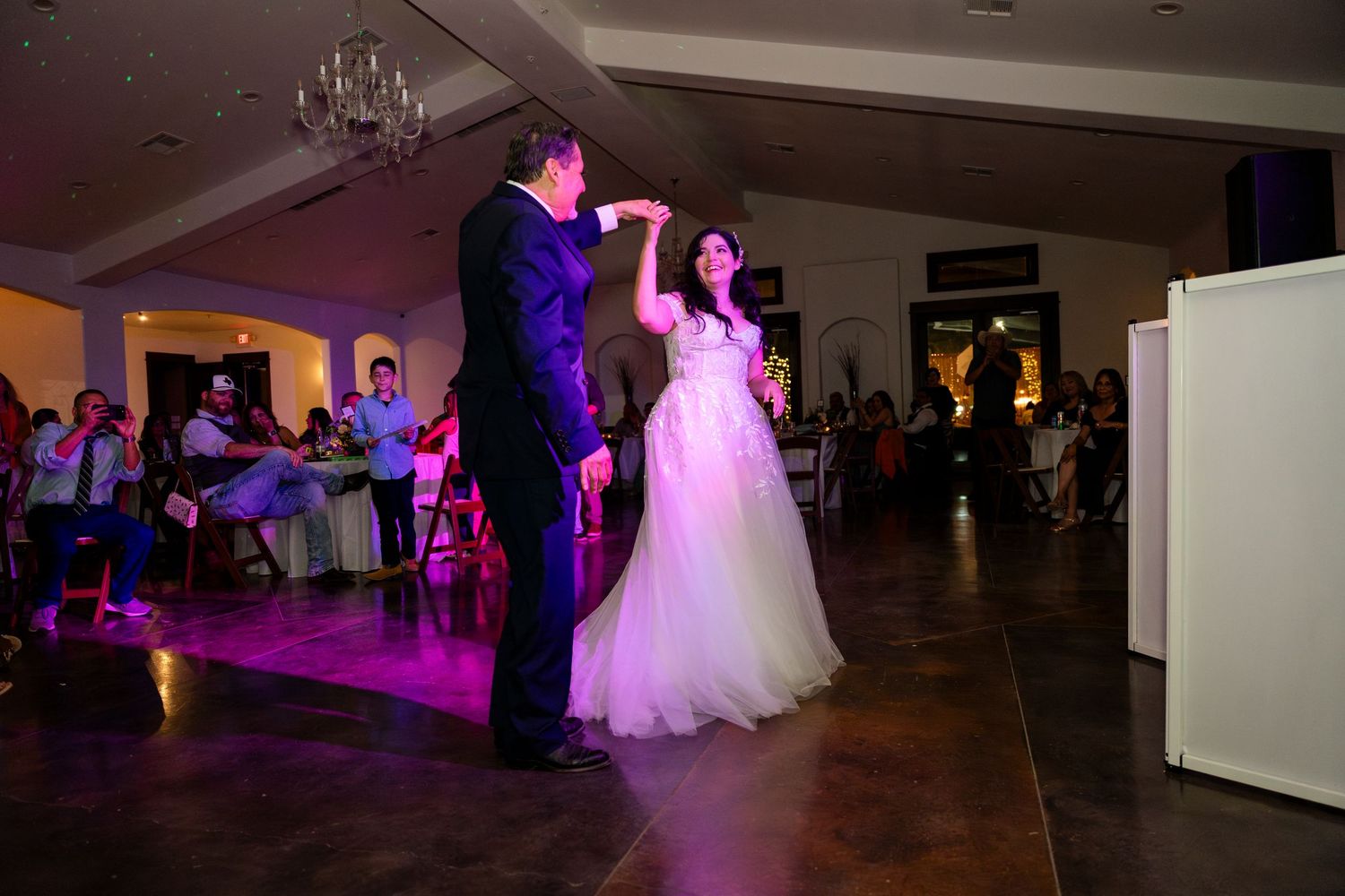 A couple shares their first dance at a wedding reception illuminated by purple lighting in an elegant ballroom.