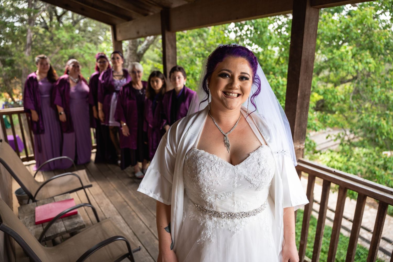 Joyful bride in white dress and purple hair poses on wooden deck with wedding party in background.