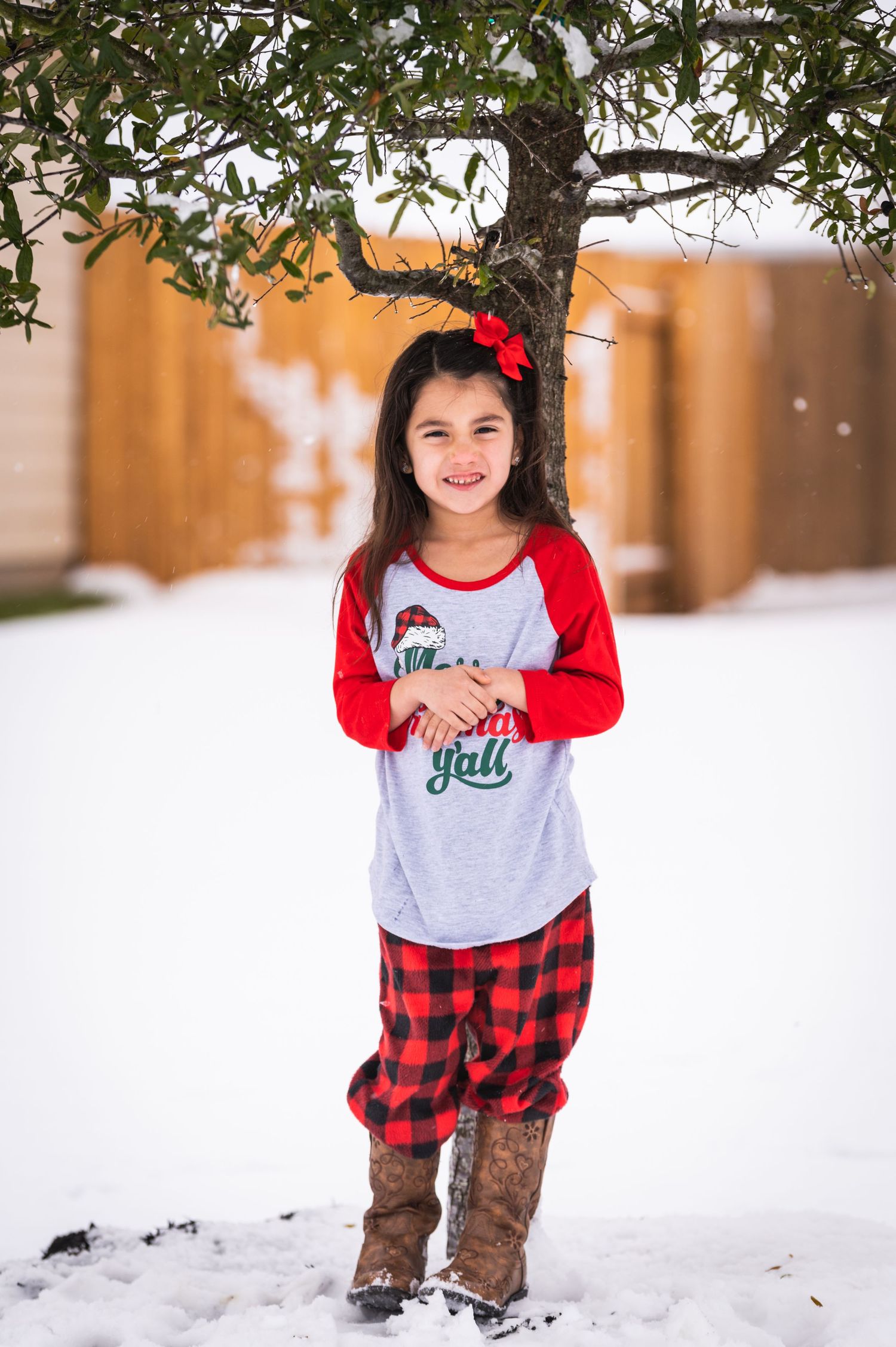 A child in holiday attire stands in snow wearing plaid pajama pants and brown boots.
