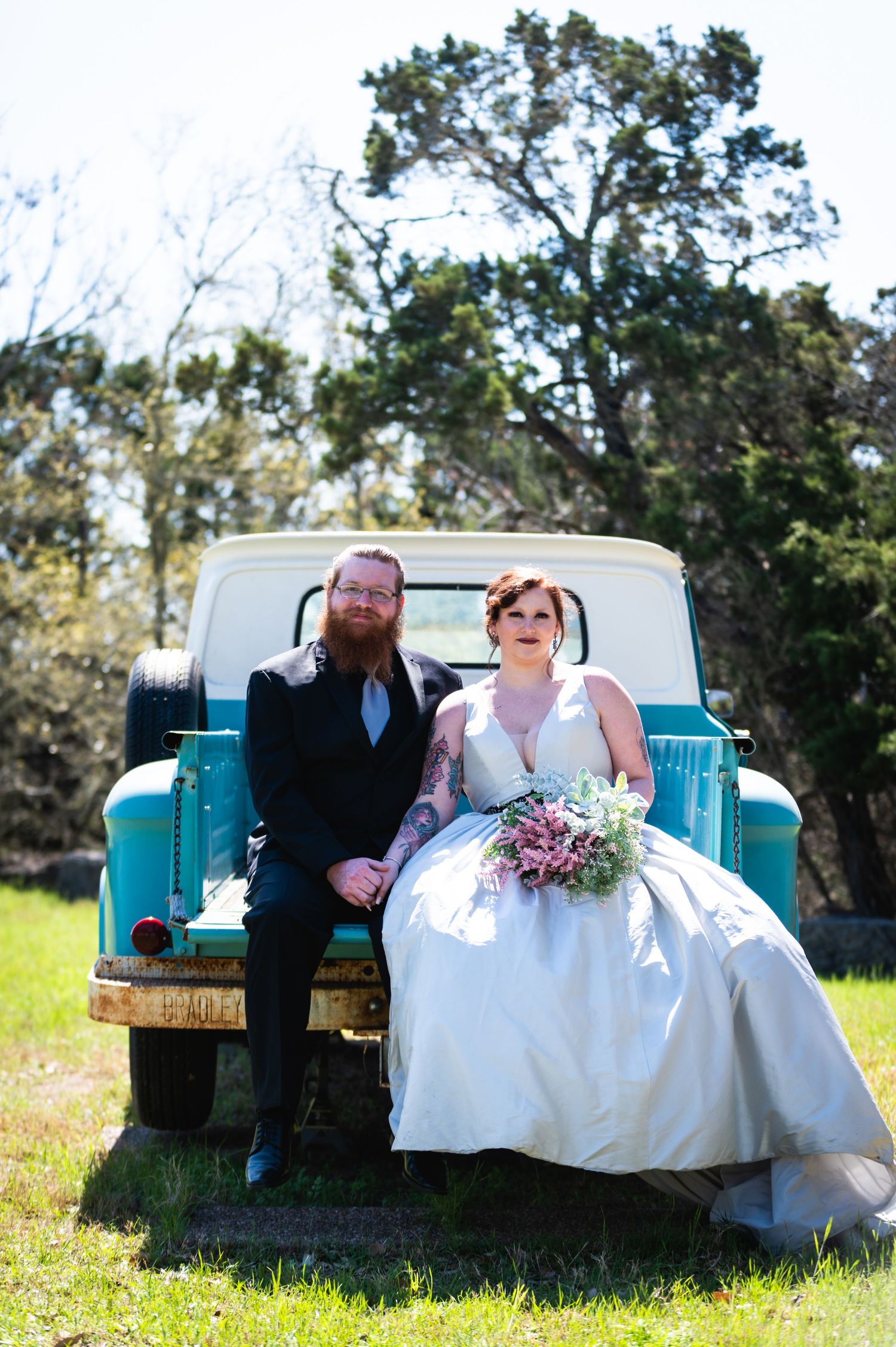 Newlyweds pose on the tailgate of a classic turquoise pickup truck outdoors.