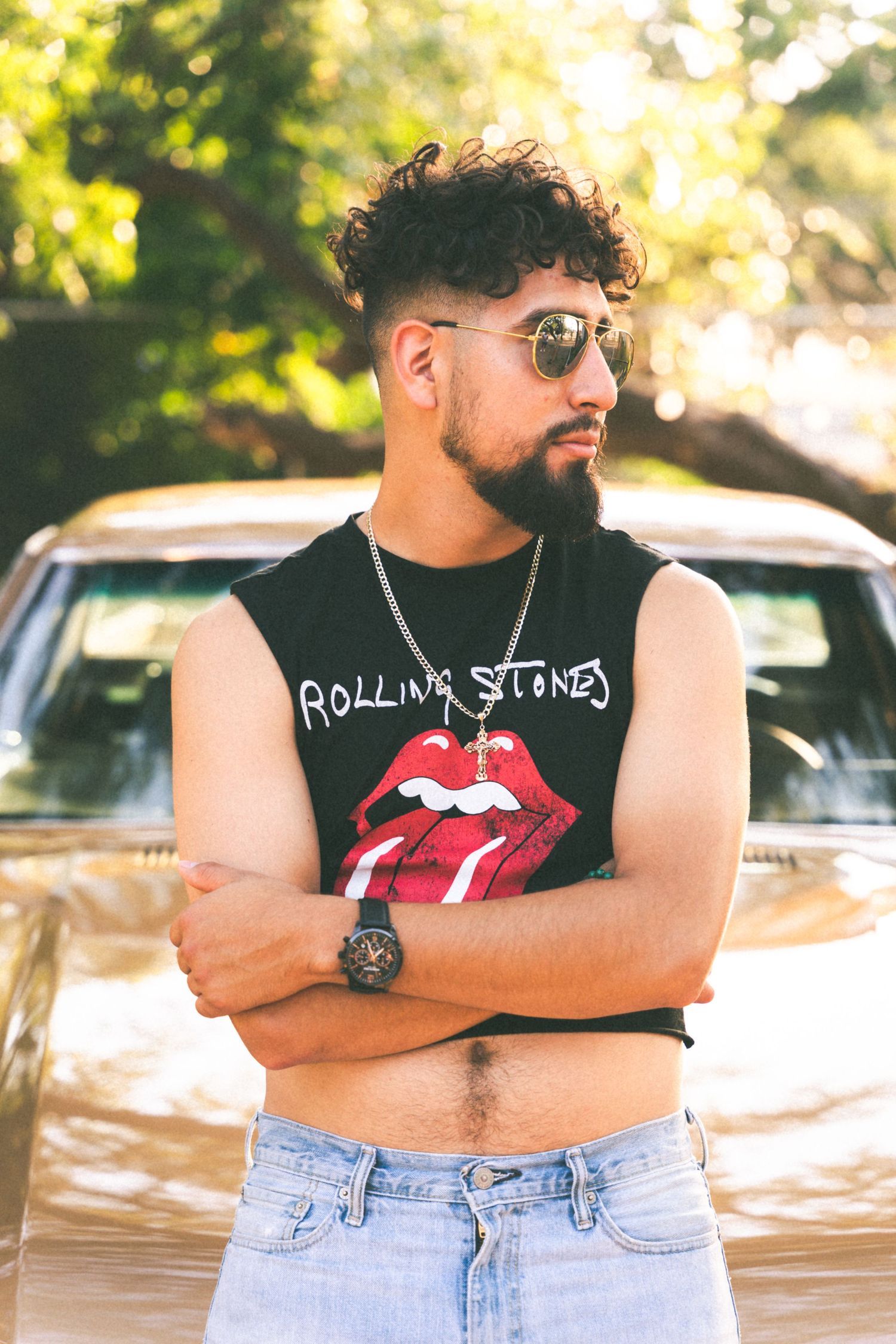 A person in a Rolling Stones tank top and sunglasses posing against a car in golden sunlight.