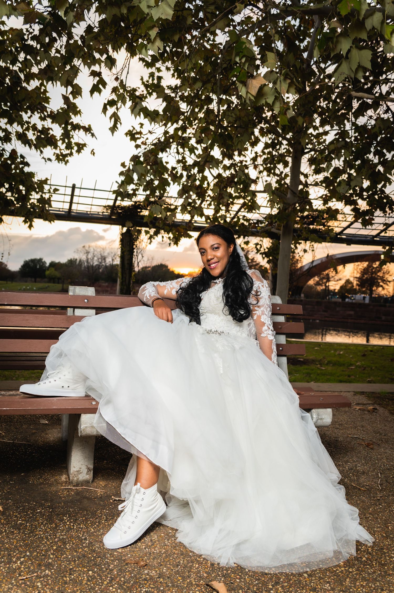 A bride in white sneakers and wedding dress sits casually on a park bench during sunset.