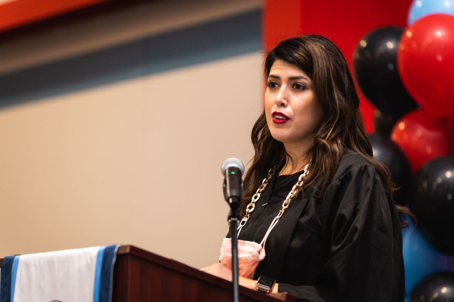 Speaker delivering remarks at graduation ceremony podium with red and black balloons in background.