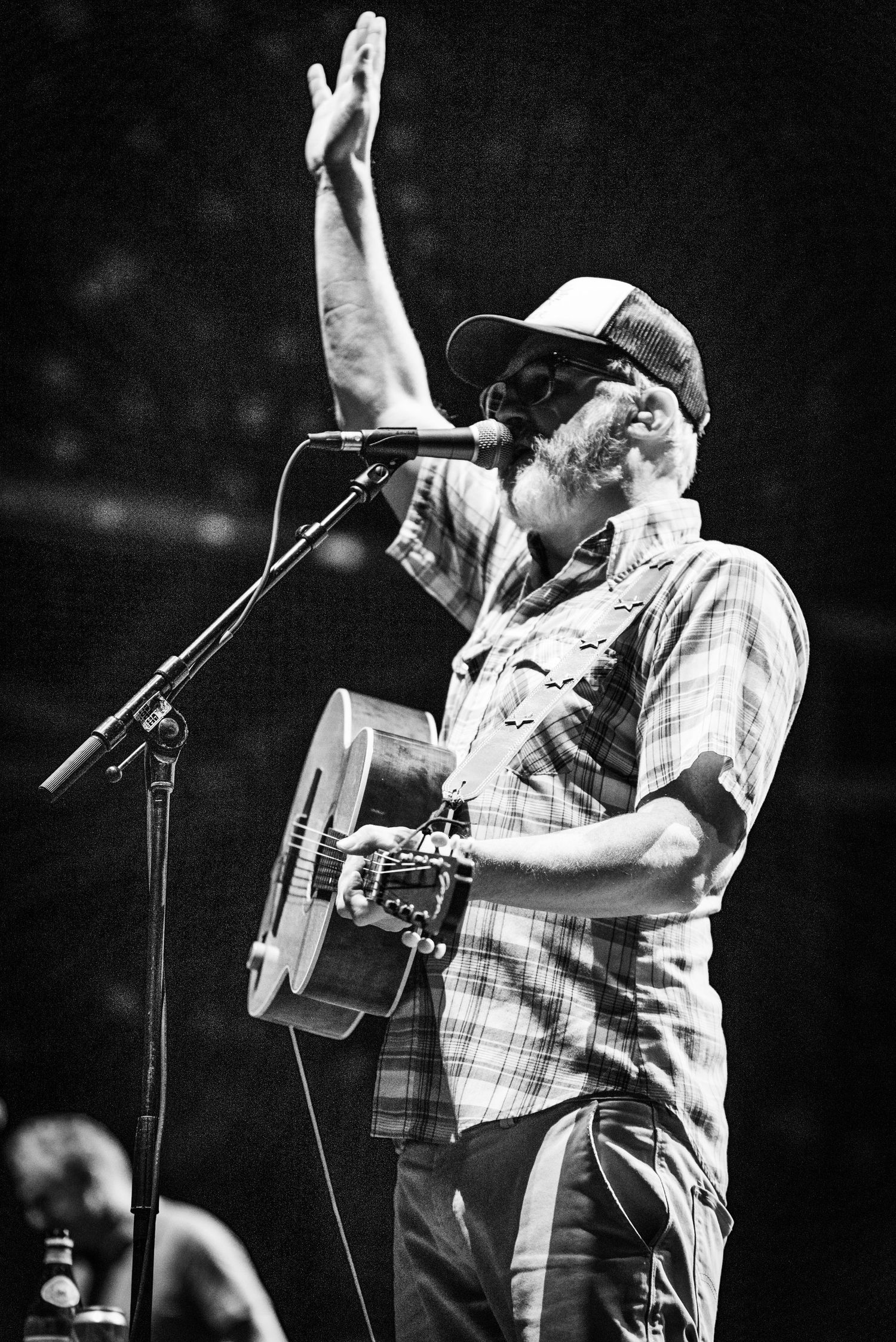 A musician in a plaid shirt and cap raises hand while performing with an acoustic guitar on stage.
