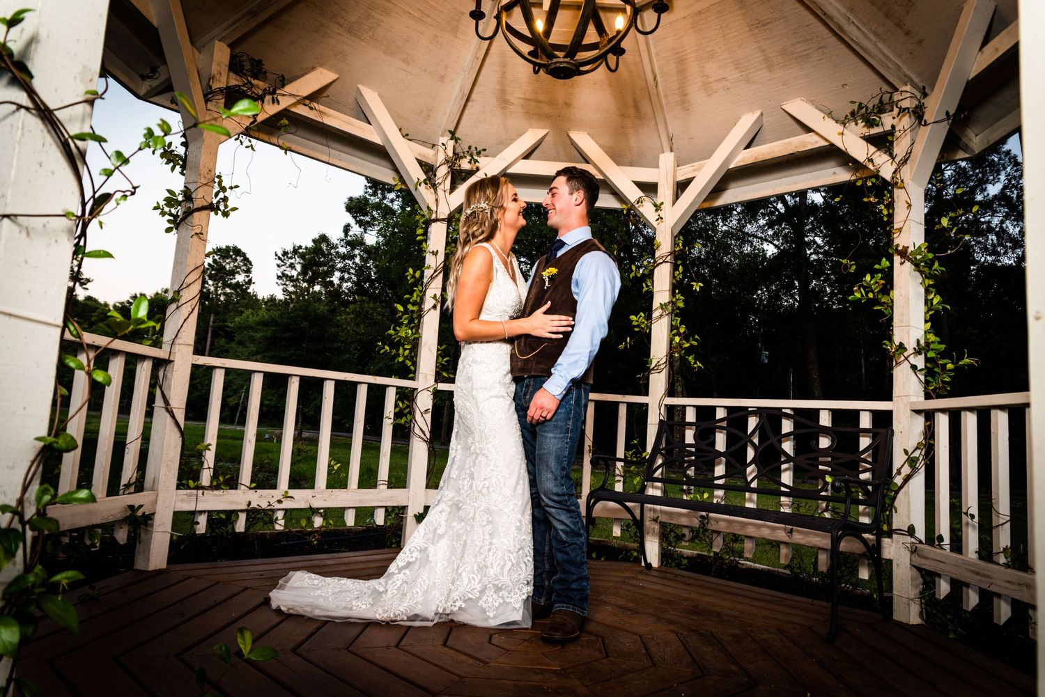 A romantic wedding couple embraces in a rustic wooden gazebo at sunset.