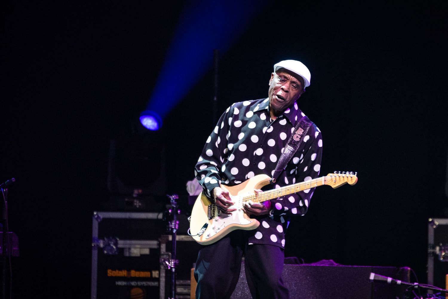Guitarist wearing polka dot shirt and cap plays Fender guitar under blue stage lights.