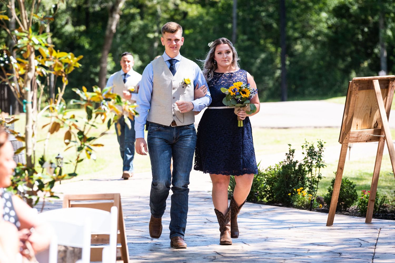 Wedding party members walk down outdoor path carrying bouquets during ceremony processional.