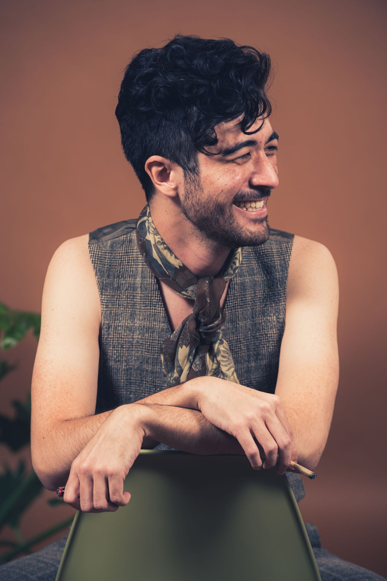 Person wearing grey vest smiles while leaning on green chair in studio with brown background.