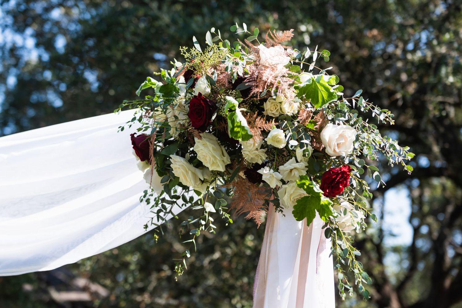 Floral wedding arch arrangement with white and burgundy flowers against sunlit greenery.