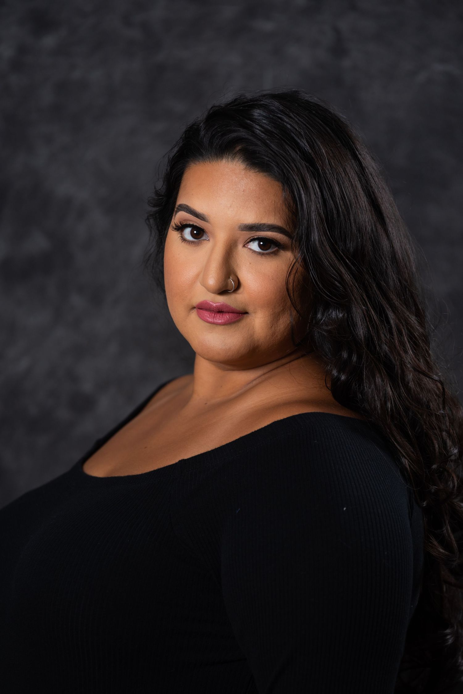 Elegant studio portrait in black attire with long wavy hair against dramatic dark backdrop.