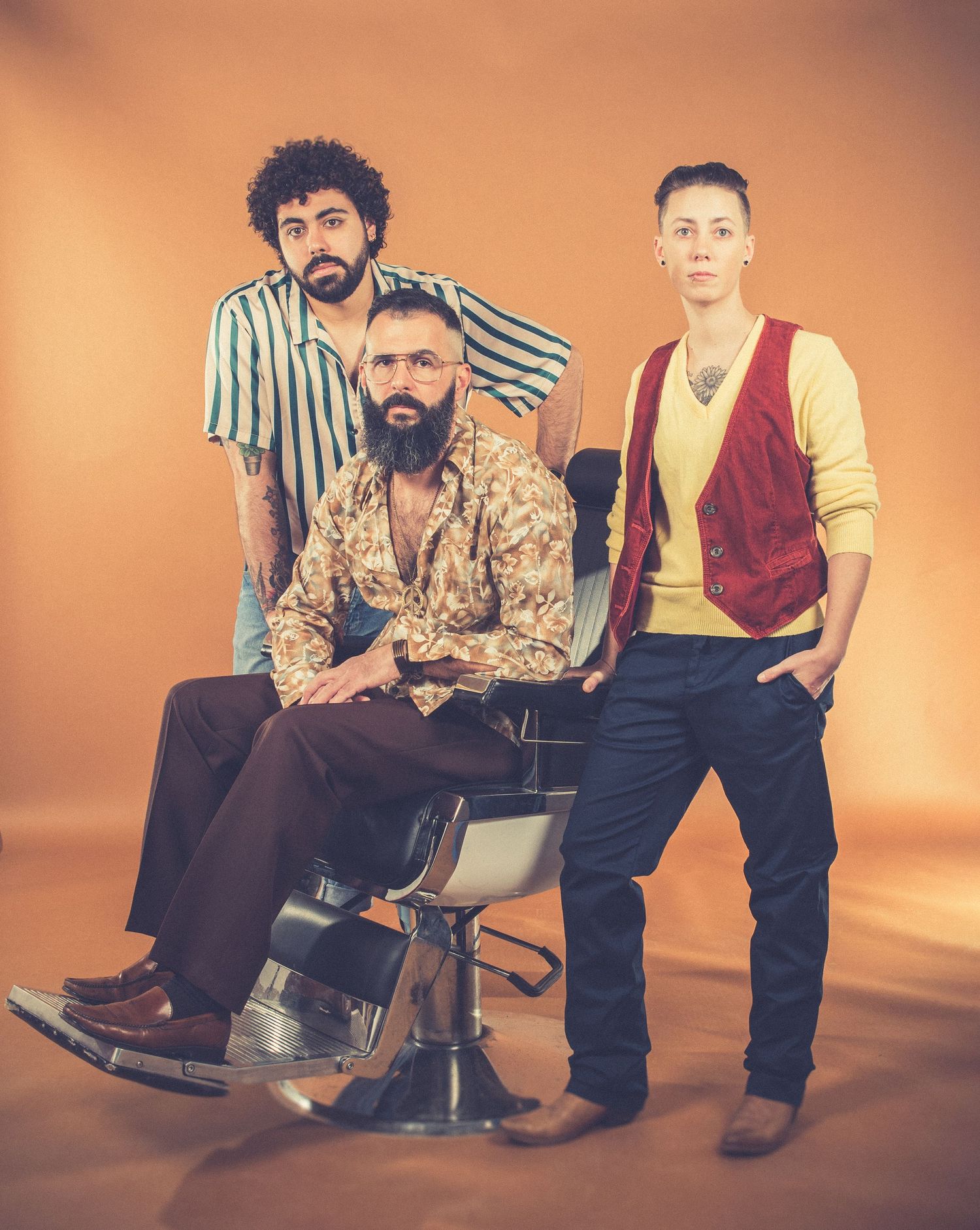 Three band members posing with one seated in a barber chair in studio setting.
