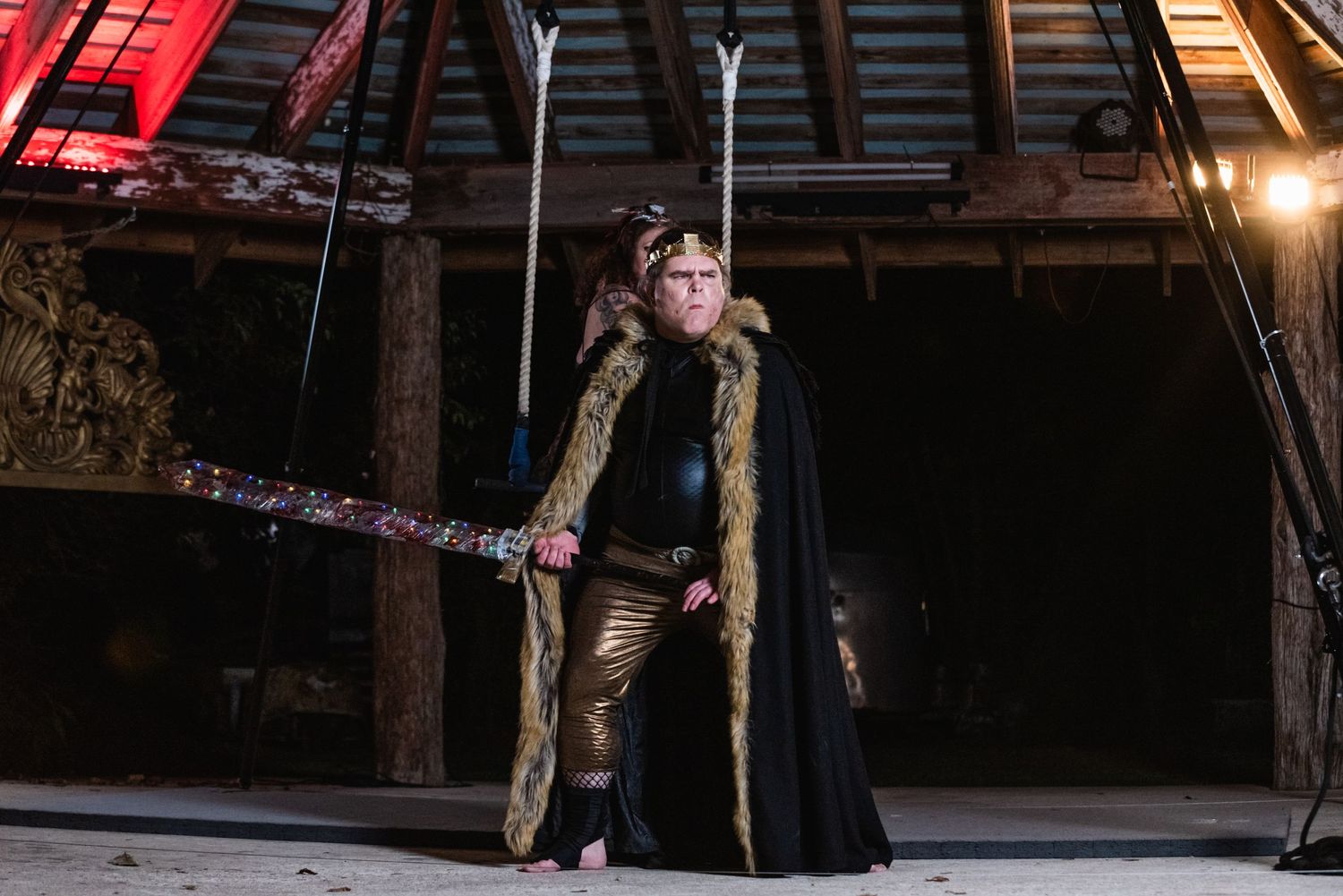 Costumed performer in black cape holds staff while standing in atmospheric wooden structure.
