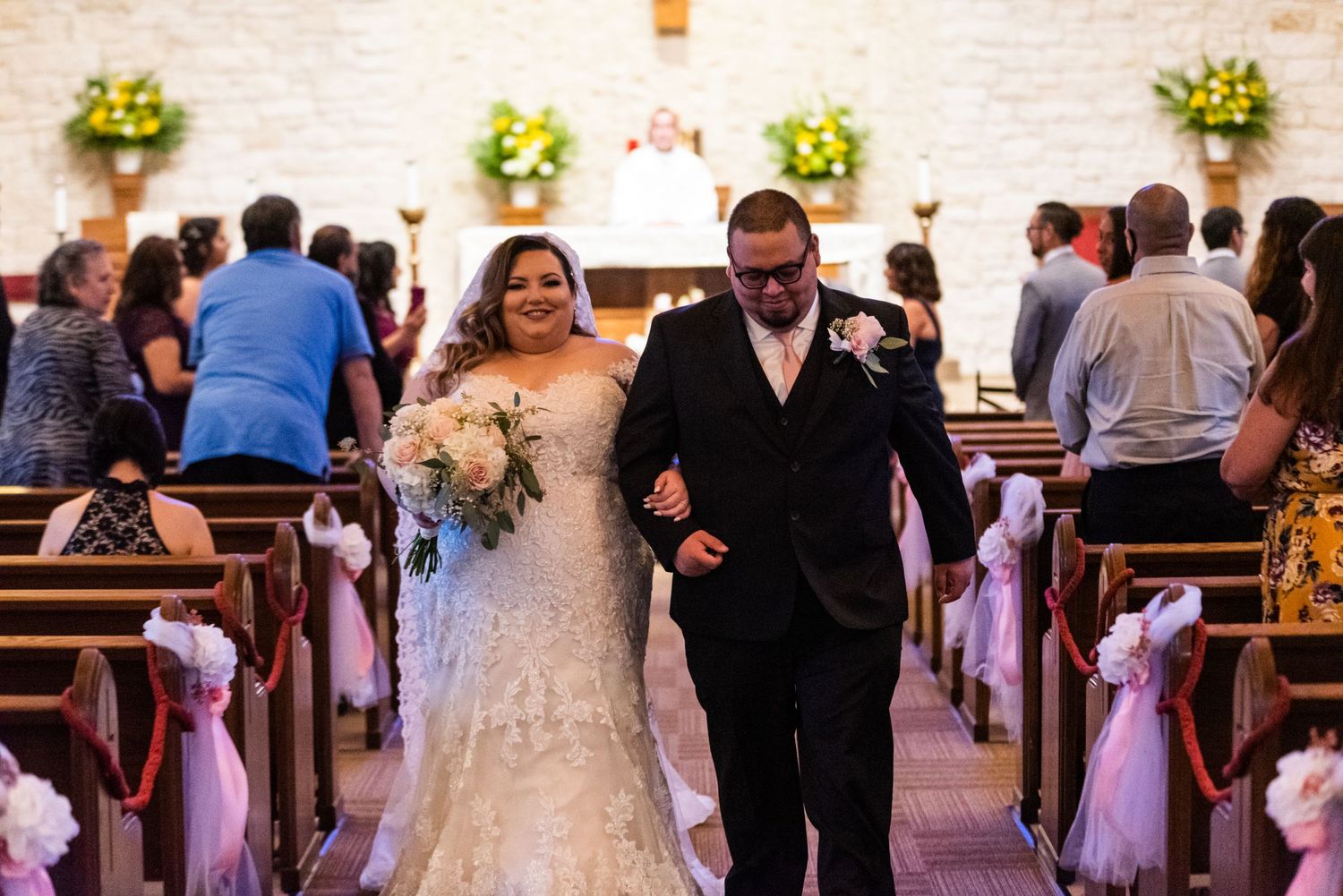 Wedding couple walks down church aisle after ceremony with floral decorations and wooden pews.