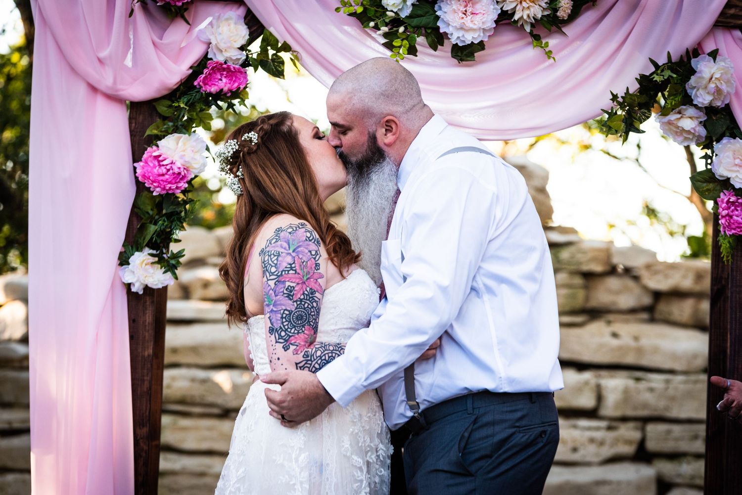 Wedding couple under pink floral arch against stone wall during ceremony.